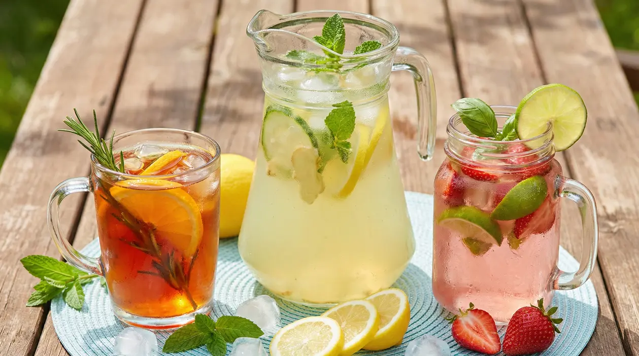 Tre bevande estive rinfrescanti con ghiaccio, agrumi, menta ed erbe aromatiche su un tavolo di legno