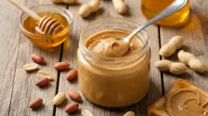 Barattolo di burro d’arachidi fatto in casa con cucchiaio, arachidi e miele su tavolo di legno