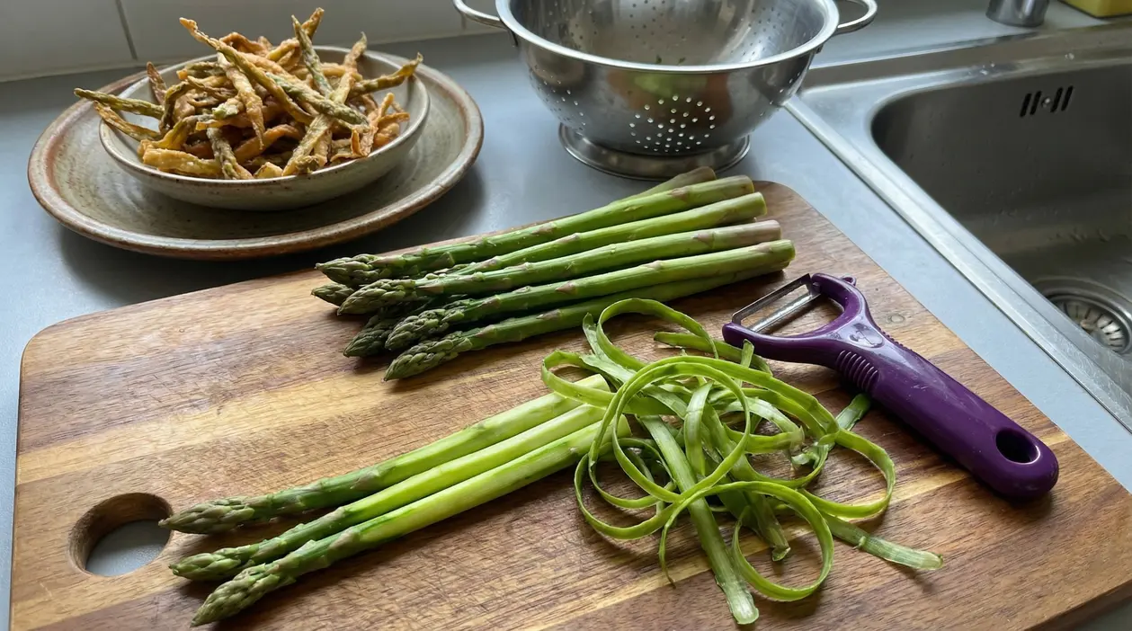 Asparagi freschi su un tagliere con bucce e pelapatate, ciotola con bucce fritte sullo sfondo